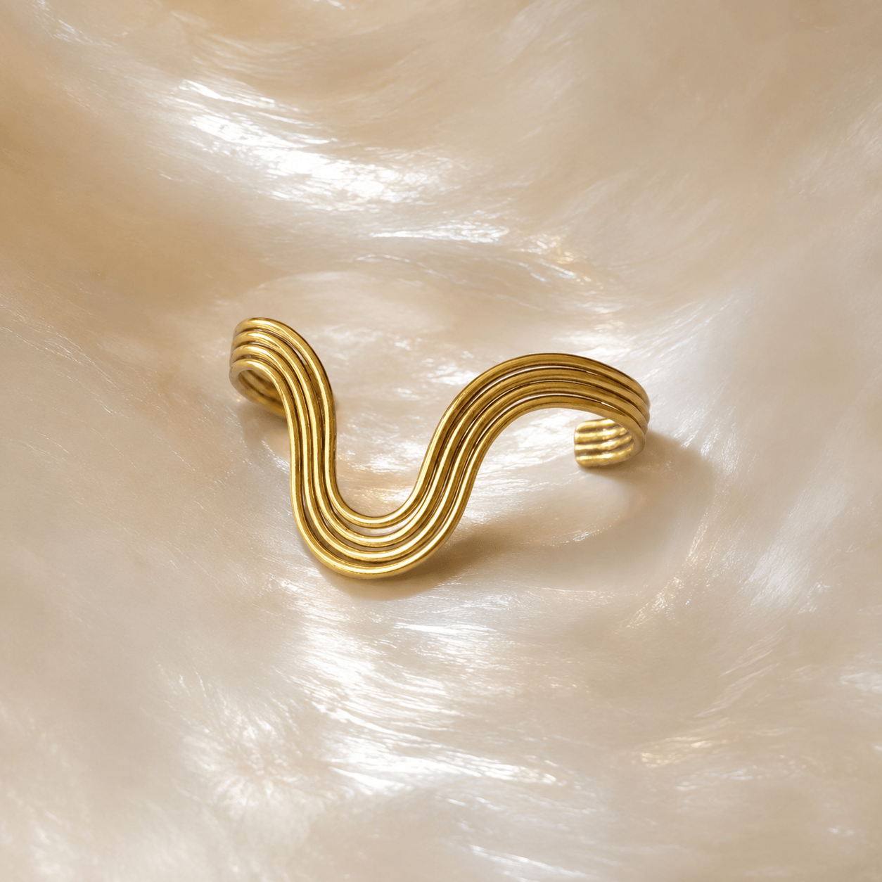 A gold-toned open cuff ring with a wavy, layered design, displayed on a textured, pearlescent cream surface.