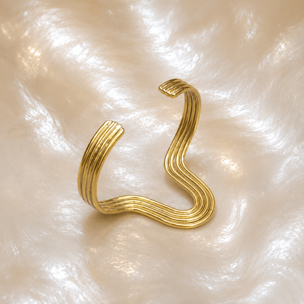 A gold-toned wavy cuff bracelet with a multi-ply ribbed design, displayed on a textured, pearlescent cream background.