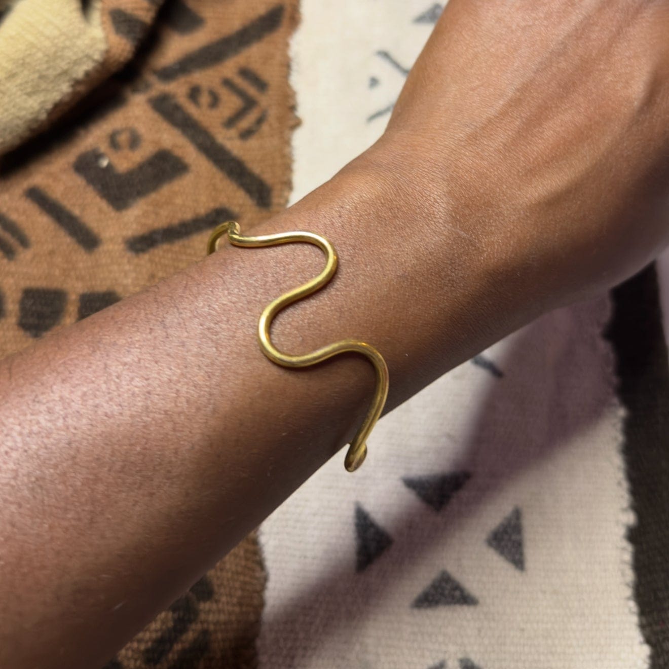 A brass wrist bracelet with a flowing curve design, worn on a person's wrist, against a patterned fabric background.