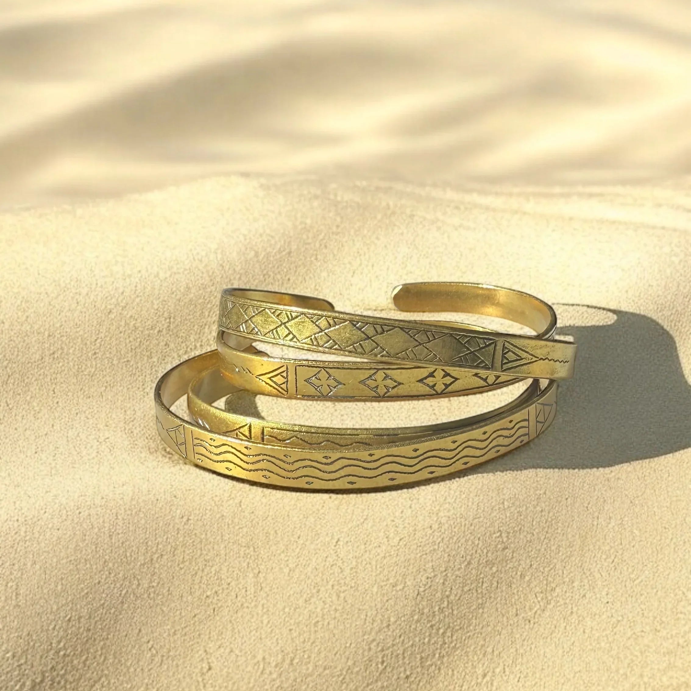 A gold-colored metal bracelet with engraved patterns, displayed on a sandy surface.