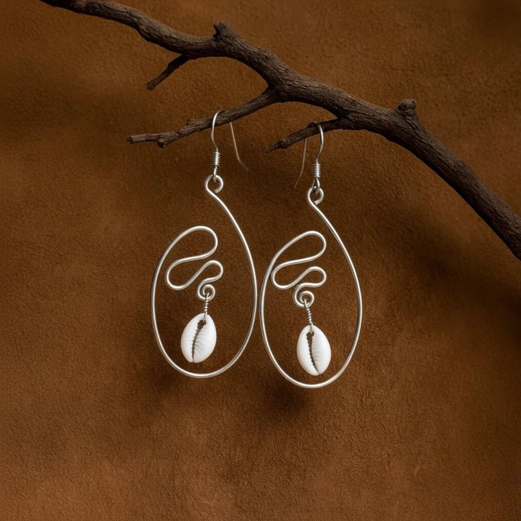 Pair of silver earrings with cowrie shells hanging on a branch against a brown background