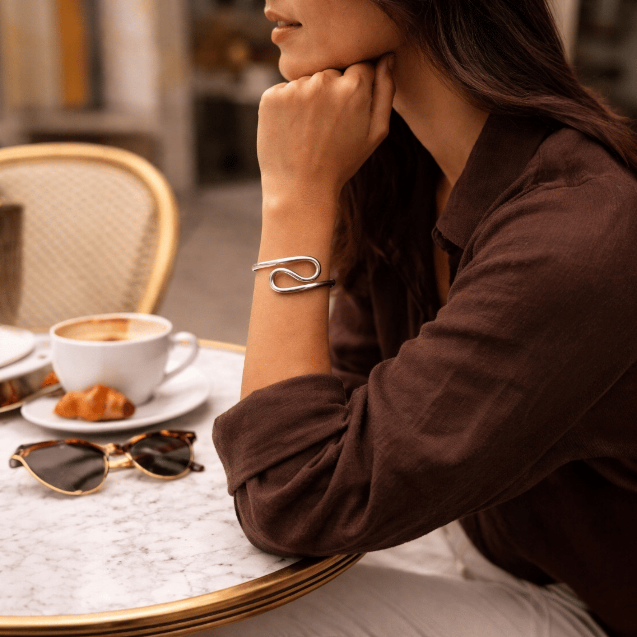 Woman sitting at a cafe table with a cup of coffee, croissant, and sunglasses.