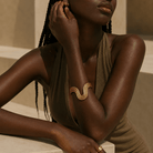 A close-up lifestyle shot of a model wearing a gold wavy cuff bracelet on her inner arm, paired with a brown halter top against a warm beige architectural background.