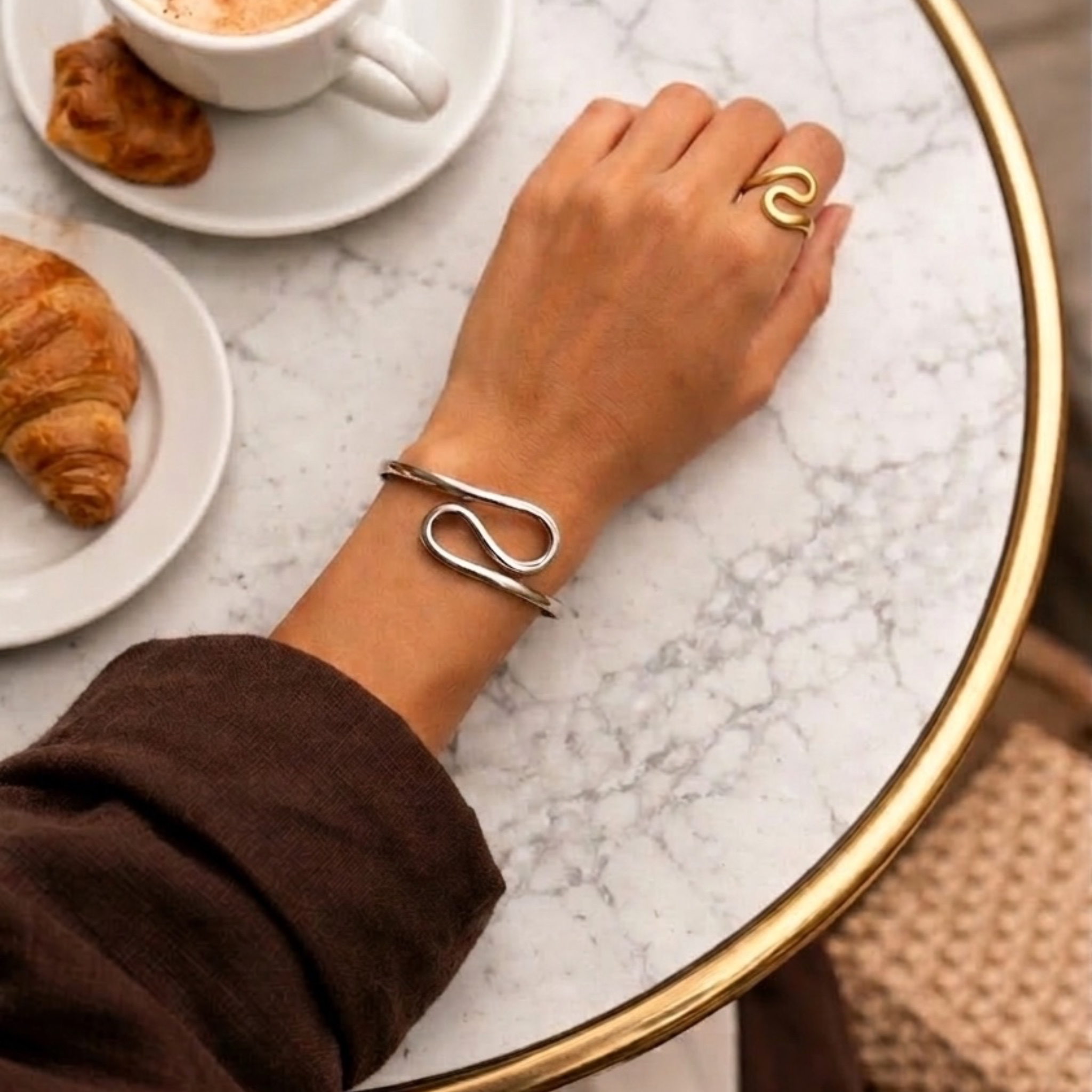 Hand wearing a silver bracelet on a marble table with a croissant and coffee.