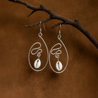 Pair of silver earrings with cowrie shells hanging on a branch against a brown background
